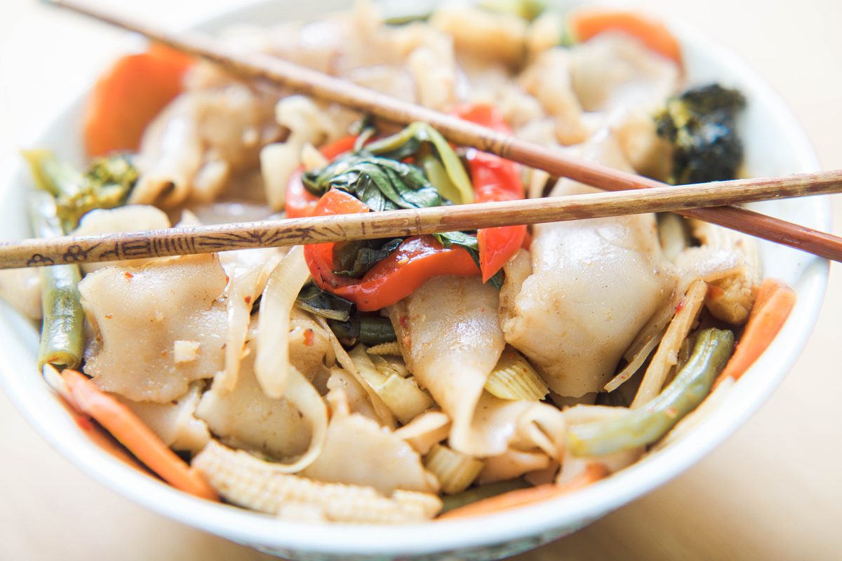 A bowl of drunken noodles with wide rice noodles, peppers, carrots and baby corn