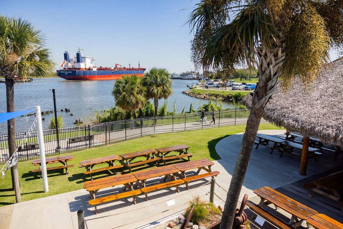 Picnic tables on the lawn at Neches River Wheelhouse in Port Neches, with a tanker passing on the Neches River