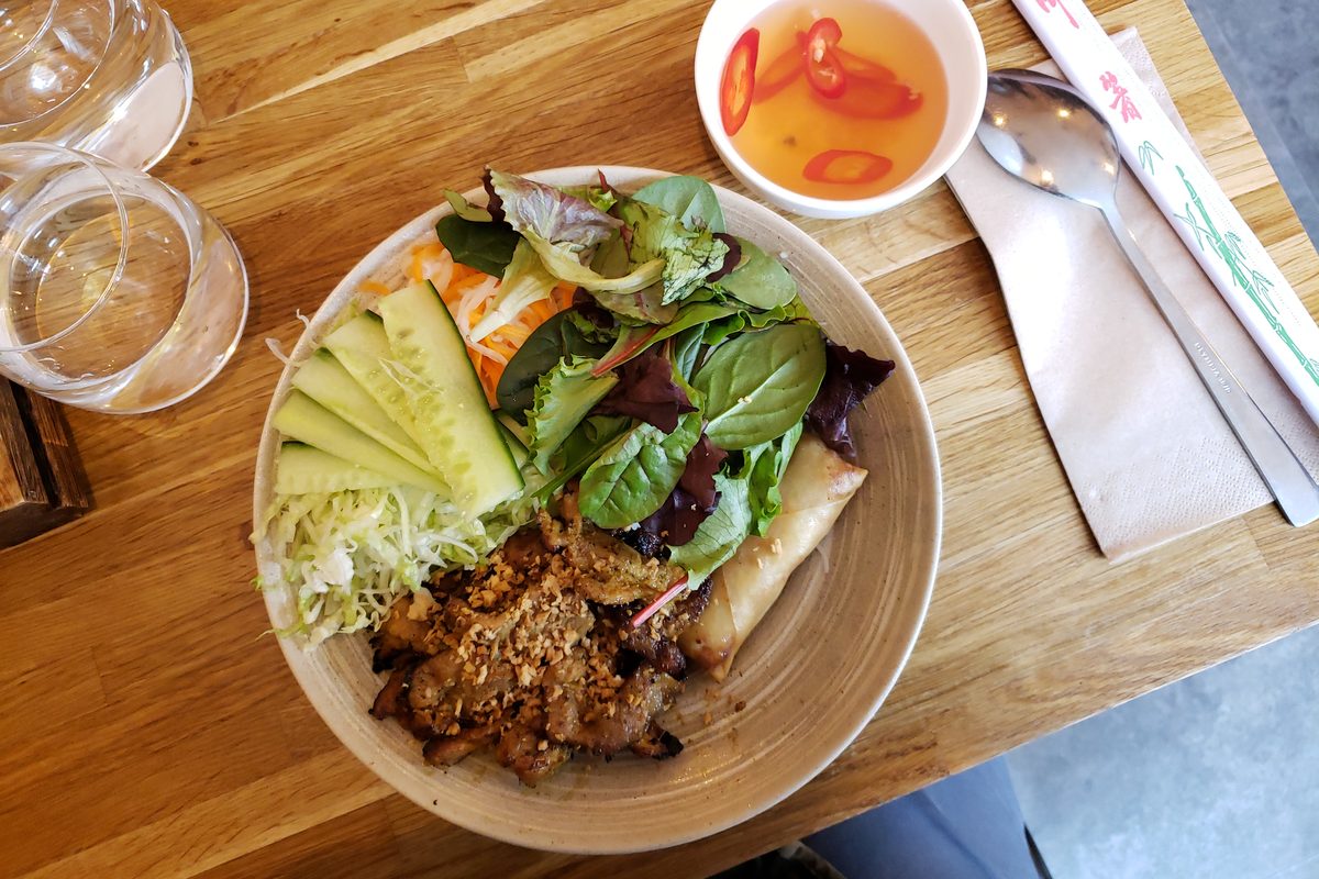 A vermicelli bowl with grilled pork, an egg roll, cucumber and greens, with a side of dipping sauce